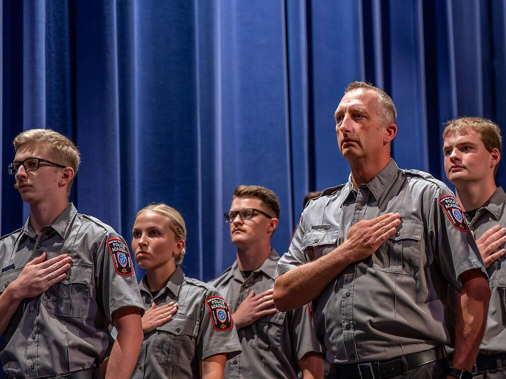 A group of uniformed Police stand in a line with hands over their hearts, facing forward, against a blue curtain backdrop.